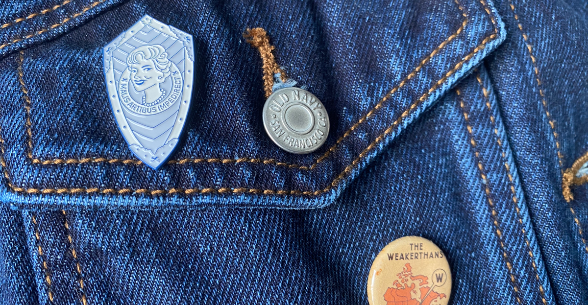 Closeup photo of the left front pocket of a blue denim jacket with an old Weakerthans button and an enamel pin that says "Malis Artibus Impedirect"
